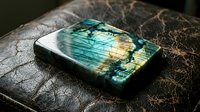 Polished labradorite gemstone with brilliant blue and green flash on textured dark surface showcasing natural iridescent mineral specimen for jewelry making.