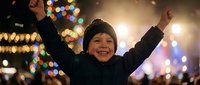 Young boy celebrating with raised arms against festive bokeh lights background during evening holiday celebration.