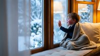 Young blonde woman in cozy sweater sitting by wooden window frame, waving goodbye or hello. Warm indoor lighting creates intimate atmosphere for lifestyle content.