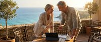 Mature Caucasian business couple working together on laptop at seaside terrace during golden hour, discussing project with ocean view background.