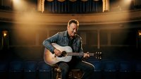Middle-aged Caucasian male musician playing acoustic guitar on stage in an empty theater, warm spotlight, casual denim style.