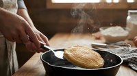 Hands flipping golden pancake in black skillet with steam rising, rustic kitchen setting with warm natural light, cooking breakfast food preparation.
