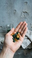 Hand holding assorted vitamin supplements and capsules in amber and green colors against weathered concrete background with glass jar for healthcare and wellness concepts.