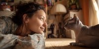 Young woman with curly hair lying on bed gazing lovingly at gray cat in warm sunlit bedroom. Peaceful morning moment between pet owner and feline companion.