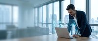 Professional businessman working on laptop in modern office conference room with city view windows during daytime meeting preparation.