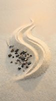 Scattered dark pebbles arranged on sandy beach with flowing wave patterns carved into sand, creating natural abstract composition for coastal and nature designs.