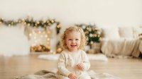 Toddler girl with curly blonde hair, approximately 1-2 years old, wearing cream-colored clothing, sitting on wooden floor with festive garland and bokeh lights in background for holiday portraits.