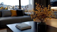 Dried golden branches in black ceramic vase on modern coffee table with books, cozy living room interior with gray sofa and soft bokeh lights in background.