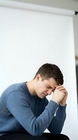 Stressed young Caucasian man in blue sweater holding head in hands showing fatigue, depression, headache or mental health struggles at workplace.