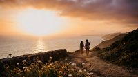 Couple walking on coastal path at sunset with golden light reflecting on ocean water and cliffs.