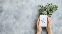Female hands holding white gift box with silver star decoration and fresh eucalyptus branches on textured grey background. Minimal holiday composition.
