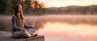 Young woman meditating peacefully on wooden dock at sunset by calm lake, practicing mindfulness and relaxation in serene natural setting for wellness.