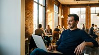 Smiling Caucasian businessman working on laptop in modern brick loft office coworking space with colleagues in background during productive workday.