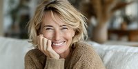 Cheerful middle-aged Caucasian woman with blonde bob haircut smiling warmly while resting chin on hands, wearing cozy beige sweater in bright living room.