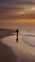Silhouetted surfer in wetsuit walking along sandy beach at golden hour sunset with ocean waves and dramatic sky, suitable for travel and lifestyle marketing.