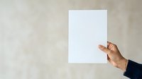 Person with medium skin tone holding blank white paper sheet against neutral textured wall background for mockup, branding, advertising, and presentation purposes.