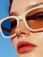 Close-up portrait of woman wearing white sunglasses with orange tinted lenses against blue sky background, showing freckled skin and coral lipstick in summer beauty style.