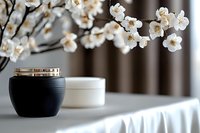 Elegant black cosmetic jar with gold lid on white table, decorated with blooming cherry blossom branch creating zen atmosphere for spa and beauty products.