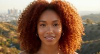 Young African American woman with curly red hair smiling outdoors with city skyline in background during golden hour sunset light for lifestyle photography.
