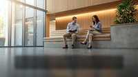Two business professionals, a man and a woman, discussing work in a modern corporate lobby with warm wooden accents and natural light.