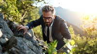 Mature businessman in suit climbing rocky mountain at sunrise, demonstrating determination and challenge in nature setting for leadership concept.