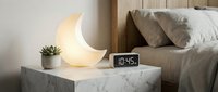 Modern bedroom nightstand with glowing crescent moon lamp, digital alarm clock and small succulent plant creating cozy ambient lighting for relaxation.