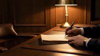 Professional businessman writing in notebook at wooden desk under warm lamp light creating focused work atmosphere for late night office tasks.