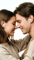 Young Caucasian couple sharing earphones, foreheads touching, eyes closed, smiling tenderly. Romantic music connection moment on light background.
