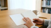Person with medium skin tone writing in spiral notebook at wooden desk with coffee cup and blurred bookshelf background, suitable for education, planning, and workspace concepts.