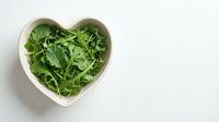 Fresh arugula leaves in heart-shaped bowl on white background. Healthy green salad ingredient for cooking and nutrition concepts.