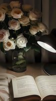 Elegant cream roses in glass vase next to open book and desk lamp creating cozy evening atmosphere. Moody lighting highlights delicate petals and reading corner.