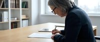 Mature woman with gray hair and glasses writing on paper at wooden desk in bright office with bookshelves, suitable for business, education, and professional concepts.