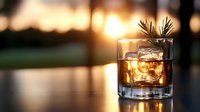 Whiskey glass with ice cubes and pine garnish against golden sunset background, creating luxurious mood for bar menu or spirits advertising.