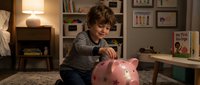 Young boy with brown hair and light skin tone, wearing a gray sweater, inserting a coin into a pink piggy bank in a cozy bedroom with warm lighting and shelving. Concept for savings, financial education, and childhood money management.