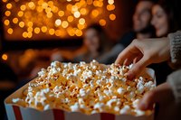 Hand reaching for popcorn in striped container with friends enjoying movie night, warm bokeh lights in background.