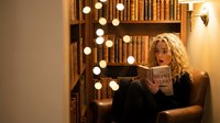 Young blonde woman reading book in cozy library corner with warm string lights and wooden bookshelves creating intimate study atmosphere for relaxation.