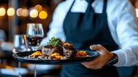 Professional waiter serving gourmet beef steak with herbs and vegetables on elegant black plate in upscale restaurant with warm ambient lighting.