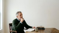 Mature man with gray hair and Caucasian ethnicity sitting at wooden desk in contemplative pose, hand to chin, wearing green sweater, surrounded by books and coffee cup, representing thoughtful professional or academic work.