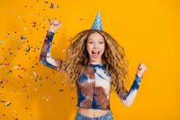 Excited young woman with curly hair celebrating birthday party with confetti falling around her on bright orange background for festive occasions.