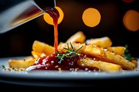 Golden french fries with rich red sauce being poured from bottle, garnished with fresh herbs and salt crystals on white plate with warm bokeh lighting.