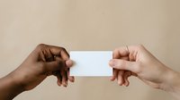 Two hands of different ethnicities holding blank white card against neutral beige background for business, networking, or promotional messaging purposes.