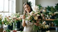 Woman florist arranging fresh cut roses and garden flowers in vase near bright window. Floral design, flower shop, wedding arrangements, event decoration, botanical styling.