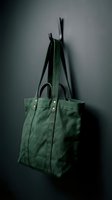 Green canvas tote bag hanging on modern black wall hook against dark gray background for fashion and lifestyle branding.