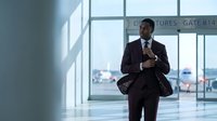 Professional African American businessman in dark suit checking smartphone while walking through modern airport terminal with large windows and departure gate signage.