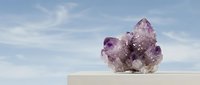 Amethyst crystal cluster on a white podium against a soft blue sky, showcasing natural purple gemstone beauty for wellness and decor.