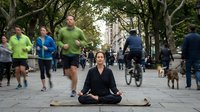 Young woman practicing meditation in lotus pose on yoga mat in busy urban park with joggers and pedestrians creating peaceful contrast in city life.