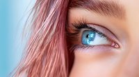 Young woman with vibrant pink hair and striking blue eyes looking upward with natural makeup and long eyelashes against soft blue background.