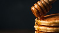Golden honey dripping from wooden dipper onto stack of pancakes against dark background for breakfast and cooking concepts.