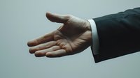 Professional businessman extending open hand in formal suit offering handshake or assistance gesture against neutral background for business concepts.