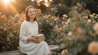 Middle-aged Caucasian woman in white dress sitting peacefully in blooming rose garden during golden hour sunset, holding coffee cup with serene expression.